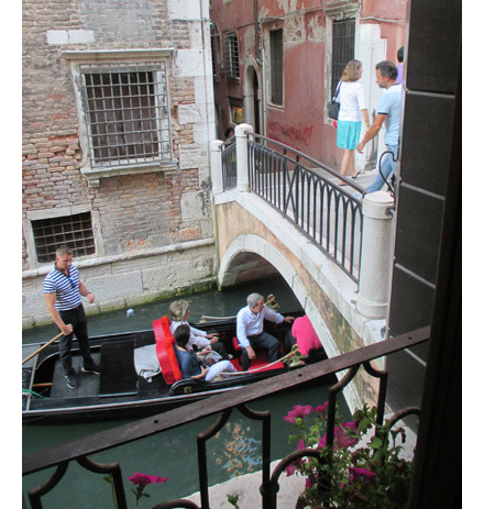 2 photos look out the second-story window onto a canal about 25 feet wide.  Each photo has a gondola with a man standing on the back of the gondola, holding the tiller and wearing black pants and a white-and-black striped shirt.  One gondola has a couple with white hair, sitting back and looking to the sides and smiling, the other has 4 people and is half-way under a small arched bridge with people standing on top of it.  The bottom of the bridge is about as high as the boatmens' chests.